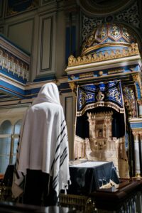 Individual in a white shawl praying at a decorative religious altar indoors.